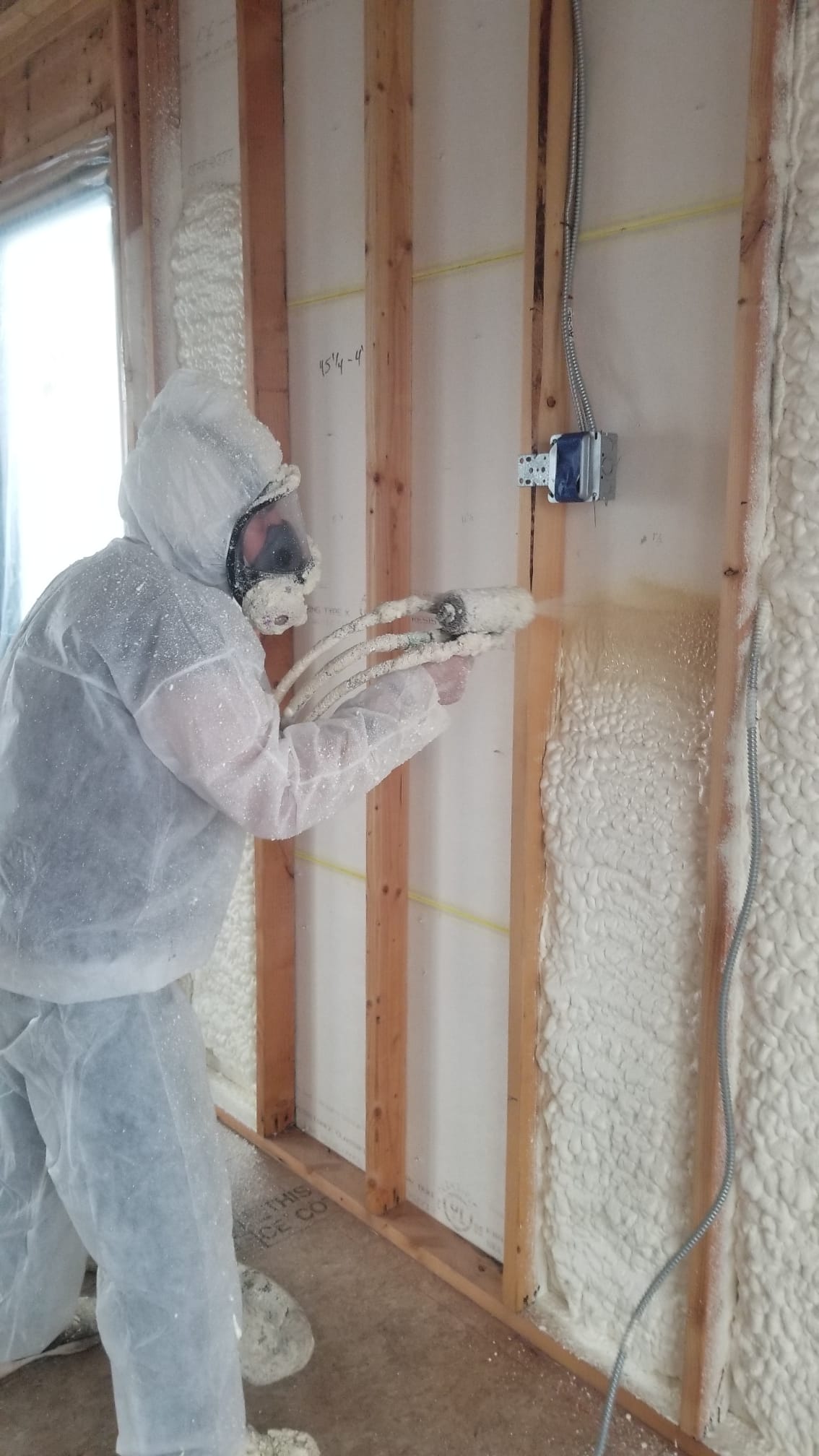 Installer in a protective suit spraying foam insulation into a framed wall